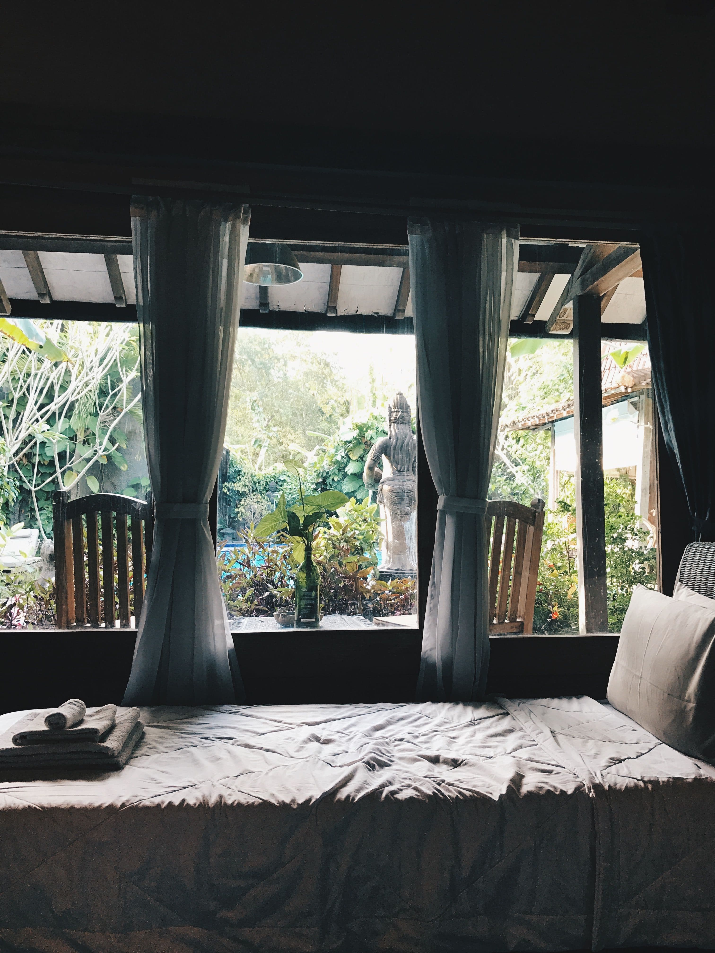 White bed and folded towels beside curtained windows overlooking a leafy garden.
